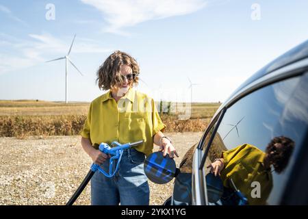 Die Frau hält eine Wasserstoff-Tankdüse. Betanken des Autos mit Wasserstoffkraftstoff. Windräder im Hintergrund. Stockfoto