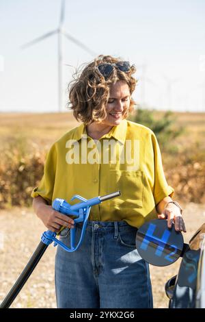 Die Frau hält eine Wasserstoff-Tankdüse. Betanken des Autos mit Wasserstoffkraftstoff. Windräder im Hintergrund. Stockfoto