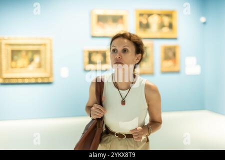 Interessierte Frau, die sich die Gemälde in der Kunstgalerie genau anschaut und den Tourbogen in der Hand hält Stockfoto