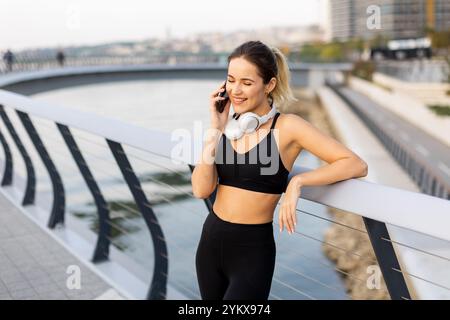 Eine Joggerin hält an ihrem Telefon am Fluss an, trägt Trainingsausrüstung und Kopfhörer und genießt die warme Sonne und die ruhige Atmosphäre Stockfoto