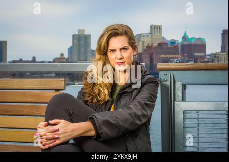 Eine Frau mit langen blonden Haaren sitzt auf einer Holzbank und trägt eine schwarze Jacke mit der Skyline der Stadt im Hintergrund. Der bewölkte Himmel erzeugt ein gedämpftes Stockfoto