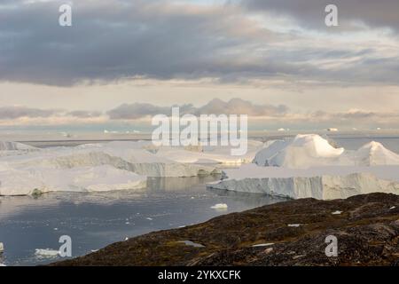 Die atemberaubende Landschaft von Ilulissat, Grönland, entfaltet sich mit einer dramatischen Mischung aus hoch aufragenden Eisbergen und zerklüfteten Felsformationen. Im Vordergrund, gezackt Stockfoto