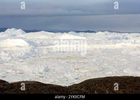 Die atemberaubende Landschaft von Ilulissat, Grönland, entfaltet sich mit einer dramatischen Mischung aus hoch aufragenden Eisbergen und zerklüfteten Felsformationen. Im Vordergrund, gezackt Stockfoto