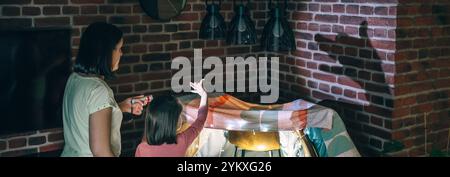 Banner der Mutter, die eine Taschenlampe hält, während die Tochter Schattenpuppen mit Händen über der Wand macht. Stockfoto