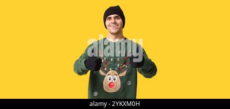 Hübscher junger Mann im Weihnachtspullover mit Daumen-oben auf gelbem Hintergrund Stockfoto