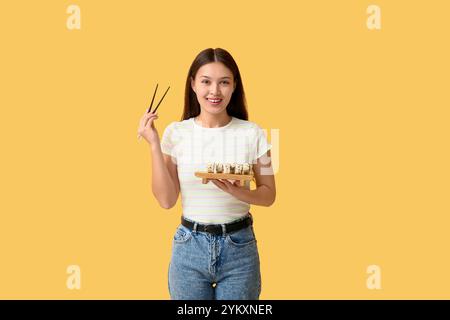 Schöne junge asiatische Frau, die leckere Sushi-Brötchen auf Holztablett und Essstäbchen auf gelbem Hintergrund hält Stockfoto