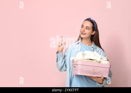 Schöne junge glückliche Frau mit Korb mit sauberer Wäsche, die auf etwas auf rosa Hintergrund zeigt Stockfoto