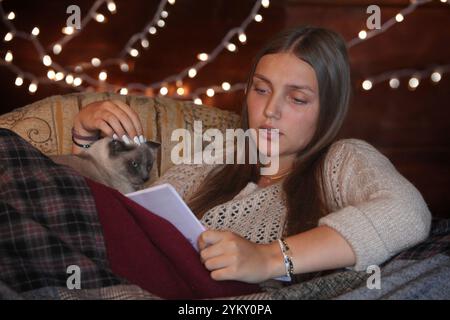 Junge Frau, die auf einer Couch sitzt, die mit einer Katze bedeckt ist und nachts ein Buch liest Stockfoto