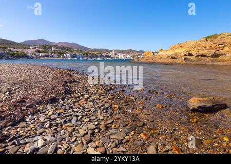 Eine mit Kieseln übersäte Küste in Cadaques, mit dem klaren Mittelmeer Wasser und dem malerischen Dorf, einschließlich der Kirche Santa Maria, im bac Stockfoto