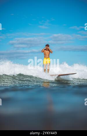Ein Mann in gelben Shorts steht auf einem Surfbrett im Meer. Er schaut in die Kamera Stockfoto