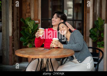 Ein junges, charmantes asiatisches Paar, das in gemütliche Winterkleidung gekleidet ist, genießt einen Kaffeedreff in einem Café in der Stadt, führt ein angenehmes Gespräch und teilt sich Stockfoto