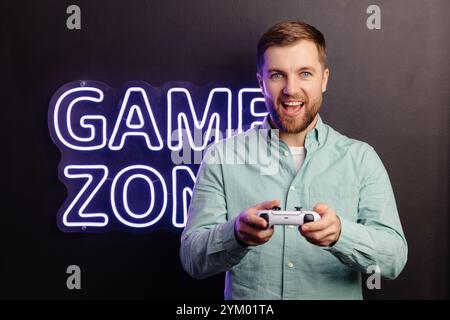 Junger bärtiger Mann, der an der Wand im Raum steht und spielt, den kabellosen Joystick hält, schreiend und ein intensives Videospiel gewinnt. Stockfoto