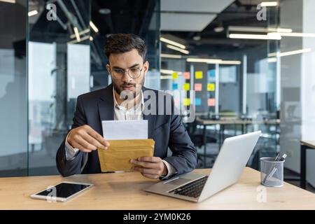Ernsthafter Mann im Amt, der Briefumschlag hält, mit dem Lesen von Nachrichten beschäftigt. In Business-Kleidung gekleidet, die Fokus und Konzentration zeigt. Technologie wie Laptop und Telefon sind vorhanden. Stockfoto