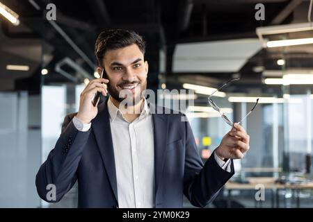 Selbstbewusster Geschäftsmann im Büro, der telefoniert, lächelt, während er eine Brille hält. Steht für modernen Unternehmensstil, Kommunikation und Professionalität. Stockfoto