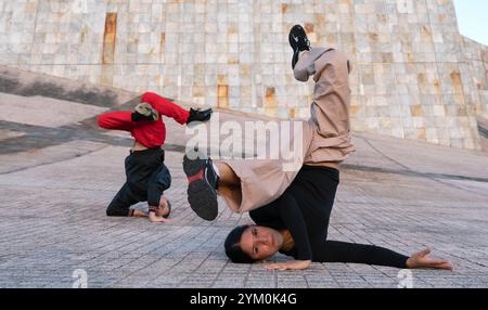 Eine Frau in einem schwarzen T-Shirt und einer braunen Hose, die eine Breakdance-Pose auf dem Boden macht. Ein Mann in roter Hose und schwarzem Sweatshirt schlägt eine andere Pose hinter sich Stockfoto