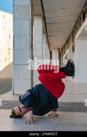 Ein junger Mann in schwarzer Jacke und roter Hose, der eine Breakdance-Pose auf dem Boden macht, seine Hände auf dem Boden liegen und die Beine eingezogen Stockfoto