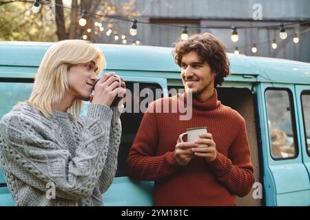 In einer bezaubernden Umgebung im Freien lachen junge schwule Paare und trinken an Tassen in der Nähe eines Vintage-Van. Ihre kuscheligen Pullover sind Empha Stockfoto