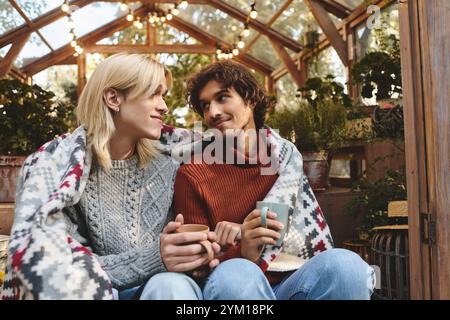 Zwei junge und gutaussehende Menschen sitzen in einer gemütlichen Decke zusammen und genießen einen friedlichen Moment in einem wunderschön beleuchteten Gewächshaus Stockfoto