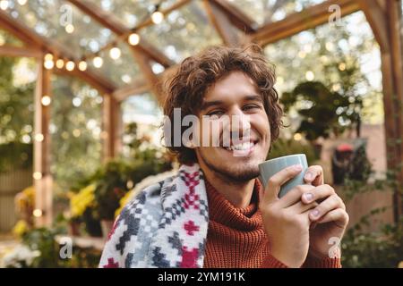 Ein junger Mann mit lockigen Haaren lächelt warm und hält eine Tasse in einem lebendigen Gewächshaus voller Pflanzen. Sonnenlicht filtert durch Glasplatten, Creat Stockfoto