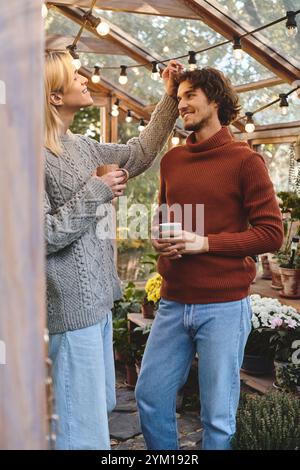 In einem ruhigen Garten voller lebendiger Blumen teilt ein junges Paar einen freudigen Moment. Das warme Leuchten der Lichterketten verstärkt die gemütliche Atmosphäre Stockfoto