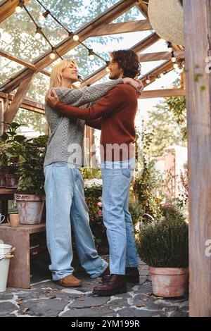 Ein hübsches junges Paar teilt einen liebevollen Moment in einem bezaubernden Gewächshaus, umgeben von üppigem Grün und gemütlichem Dekor. Sie stehen nahe und strahlen Freude aus Stockfoto