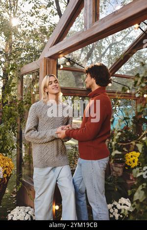 In einem bezaubernden Gewächshaus mit leuchtenden Blumen teilt ein junges Paar eine freudige Verbindung. In stylischen Pullover gekleidet, halten sie die Hände und strahlen aus Stockfoto