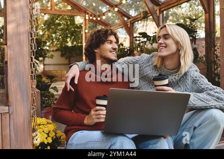 In einem bezaubernden Gewächshaus voller Grün und Blumen teilen sich das junge schwule Paar einen Moment der Freude beim Kaffeetrinken. Stockfoto