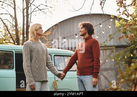 Ein junges, liebevolles schwules Paar teilt einen zärtlichen Moment, Hände miteinander verflochten, vor einem charmanten Vintage-Van-Hintergrund. Sanfte Beleuchtung und Herbstlaub sorgen für eine Wa Stockfoto