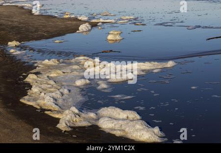 Sanfte Wellen bringen Schaum an die Sandküste, wenn die Sonne untergeht, und schaffen eine ruhige Küstenatmosphäre. Stockfoto