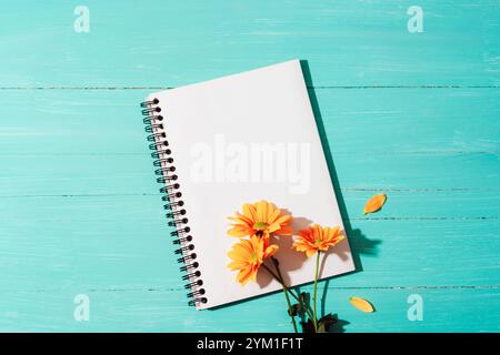 Leerer Notizblock und gelbe Blumen auf türkisblauem Holztisch mit Licht und Schatten. Draufsicht, flach, Modell Stockfoto