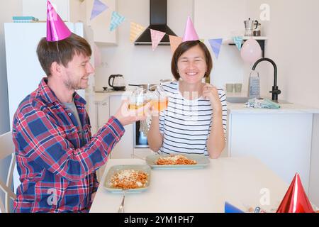Das junge Paar feiert zu Hause gemütlich Geburtstag und genießt Kuchen und Lachen in warmer Atmosphäre. Die Familie isst Pasta und trinkt Wein, um zu feiern, zu essen und zu jubeln Stockfoto