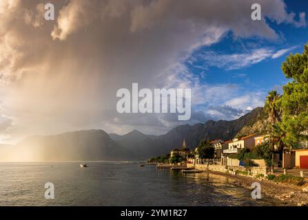 Blick auf Dobrota bei Sonnenuntergang in Kotor, Montenegro. Auf dem Foto werden ruhiges Wasser entlang der Küste, eine Reihe von Gebäuden mit üppigem Grün und eine Kulisse aus aufgenommen Stockfoto