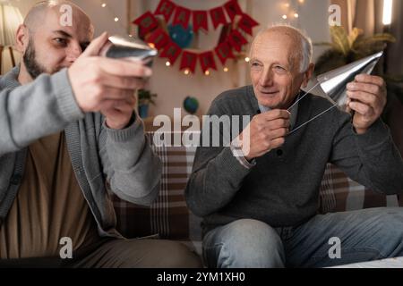 Seniorenvater und erwachsener Sohn bereiten sich vor, Geburtstag oder Jahrestag zu feiern und legen Partyhüte auf den Köpfen auf dem Sofa im Wohnzimmer Stockfoto