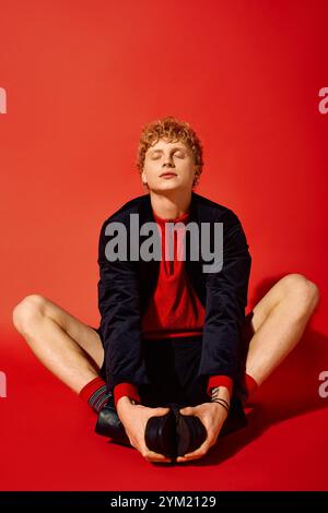Ein stilvoller Mann sitzt auf dem Boden und dehnt sich bequem in einer leuchtend roten Umgebung. Seine entspannte Pose und das trendige Outfit betonen den Geist von Christm Stockfoto