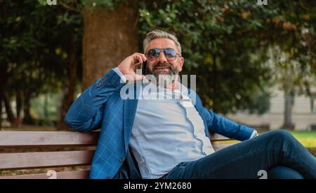 Ein glücklicher, gutaussehender, reifer Geschäftsmann, der auf dem Handy spricht, während er auf der Parkbank sitzt. . Hochwertige Fotos Stockfoto