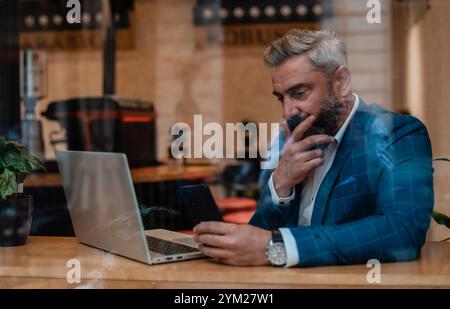 Ein durchdachter, reifer Geschäftsmann, der über eine Problemlösung nachdenkt, während er im Büro neben dem Fenster an einem Laptop arbeitet. Stockfoto
