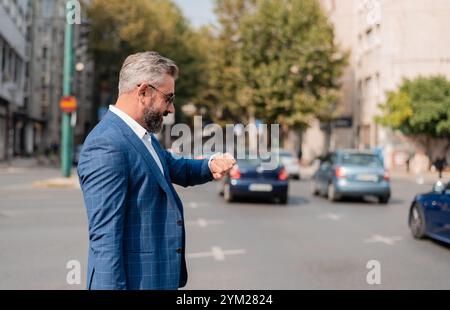 Ein geschäftiger reifer Geschäftsmann, der auf eine Armbanduhr blickt, mit Stadtverkehr im Hintergrund. Stockfoto