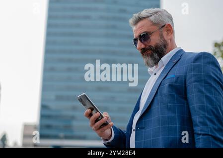 Ein gutaussehender, reifer Geschäftsmann, der außerhalb des Büros in der Stadt SMS-Apps verwendet, mit städtischen Gebäuden im Hintergrund. Stockfoto