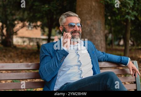 Ein glücklicher, gutaussehender, reifer Geschäftsmann, der auf dem Handy spricht, während er auf der Parkbank sitzt. . Hochwertige Fotos Stockfoto