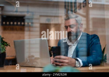 Ein gutaussehender reifer Geschäftsmann, der sein Handy benutzt, während er im Büro neben dem Fenster an einem Laptop arbeitet. Stockfoto