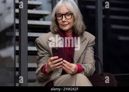 Eine stilvolle ältere Frau verwendet ihr Smartphone, während sie draußen auf einer Stufe sitzt Stockfoto