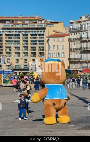 Marseille. Frankreich - 20. November 2024: Ein Kind interagiert freudig mit einem großen Maskottchen im belebten Alten Hafen von Marseille, umgeben von Menschen und Stockfoto