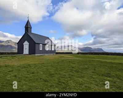 Isolierte schwarze Kirche auf Grasland mit Bergen im Hintergrund unter bewölktem Himmel. Island Stockfoto