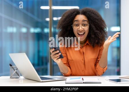 Junge afroamerikanische Geschäftsfrau im Büro, die ihr Smartphone hält und begeistert aussieht, während sie mit Laptop und Dokumenten arbeitet. Positives und energiegeladenes Arbeitsumfeld. Stockfoto