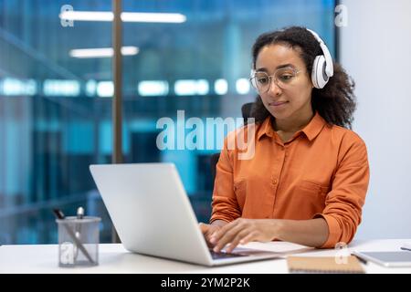 Junge afroamerikanische Geschäftsfrau, die in einem modernen Büro am Laptop arbeitet. Mit Kopfhörern wirkt sie konzentriert und engagiert. Ideal für die Themen Technologie, Geschäft und Produktivität. Stockfoto