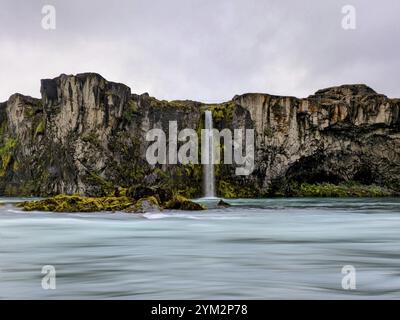 Lebendiger Wasserfall, der unter einem bewölkten Himmel moosbedeckte Klippen hinunterstürzt, isländische Landschaft. Island Stockfoto