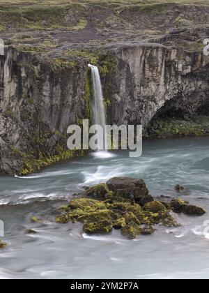 Ein ruhiger Wasserfall, der über eine felsige Klippe mit üppigem Moos in türkisfarbenes Wasser in einem Canyon stürzt. Island Stockfoto