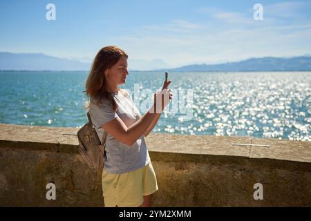 Frau macht an einem sonnigen Tag mit Blick auf die majestätischen Berge malerische Fotos an einem glitzernden See. Idyllische Lage am Seeufer bietet natürliche Schönheit und O Stockfoto