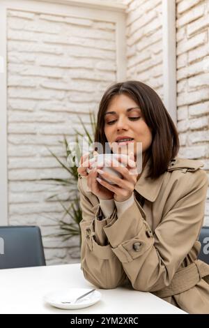 Die junge Frau in einem beigefarbenen Trenchcoat hält eine warme Tasse und genießt einen ruhigen Moment in einem Café im Freien. Neutraler Hintergrund und sanfte Beleuchtung schaffen eine Rela Stockfoto