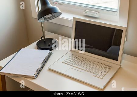 Ein Laptop steht auf einem Schreibtisch neben einem Notebook offen. Das Notebook ist schwarz-weiß und das Notebook weiß. Der Schreibtisch ist sauber und organisiert Stockfoto